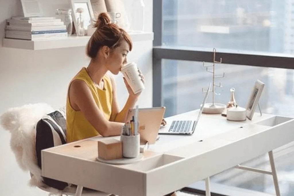 Mujer joven con blusa amarilla en oficina moderna bebiendo café y trabajando en laptop frente a ventana grande